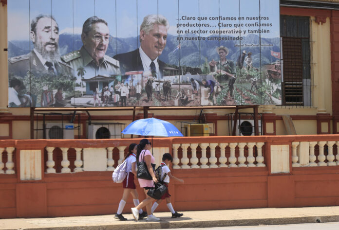 Una mujer y dos niños bajo una sombrilla fueron captados el pasado 1 de junio al caminar frente a una valla con las imágenes del presidente de Cuba, Miguel Diaz-Canel (d), y de los expresidentes Raúl (c) y Fidel Castro, en La Habana (Cuba). EFE/Ernesto Mastrascusa

