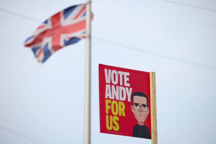 Una bandera del Reino Unido ondea cerca de un cartel del candidato laborista Andy Burnham en Wigan, Reino Unido, en una imagen del 15 de junio de 2026. La circunscripción inglesa de Makerfield celebra este jueves una elección parcial para elegir a su nuevo diputado, cita en la que Burnham concurre como candidato laborista a fin de entrar en el Parlamento y desafiar eventualmente el liderazgo de Keir Starmer. EFE/EPA/ADAM VAUGHAN
